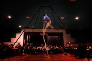 Woman performing aerial silks on white fabric in a dimly lit gymnasium at a masquerade ball