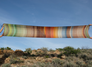 Two people holding up a large brown, red, orange, yellow, blue and green piece of handwoven fabric in the desert. 