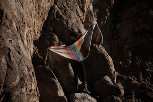 woman wearing all black and a rainbow colored scarf shawl, sitting on rocks at the base of a cliff
