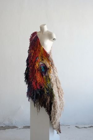 A rainbow shawl completely covered in fringe on a white mannequin, on a white pedestal, in a white room