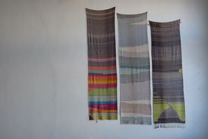 Three large woven tapestries hang on a white wall.  One is rainbow and grey, one is blue-grey one is chartreuse and grey.