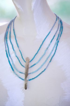 A 4 strand pale blue beaded necklace with a thin crystal centerpiece is on a white painted mannequin 