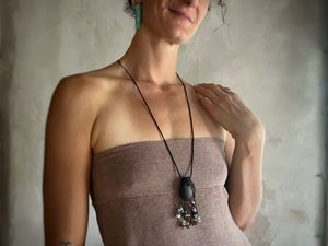 A woman wearing a pale purple top and long stone talisman necklace with dangling beads stands in front of a white mud wall