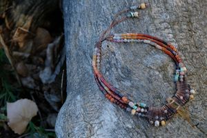 A handmade necklace of red, green, orange, purple precious stones and knotwork lay on a gray log