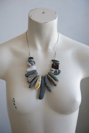A white colored female form mannequin wears a sculptural necklace of gray braided twine with rough stones hanging from it.