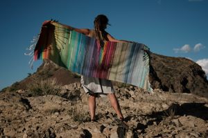 A woman wearing a many rainbow colored shawl stands in bright desert light on a Rocky Mountain 