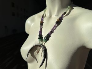 A white mannequin wears a necklace with a silver crescent, moon and green and purple, semi precious stone beads