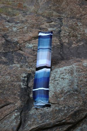 A large piece of handwoven fabric in shades of white to blue to black floats on a large rock cliff