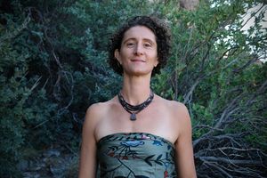 A woman wears a necklace of dark shell beads against a dark wooded background 