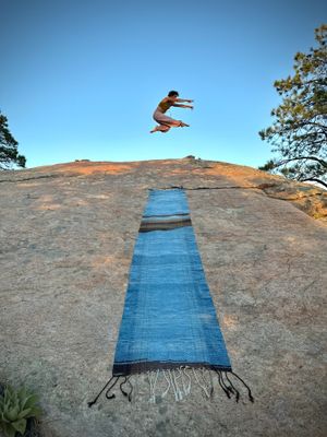 a woman jumping on the horizon with an indigo dyed blanket laying on a rock in the foreground