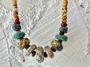 A necklace of Gray white and yellow granite beads hangs against the white wall 