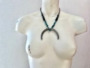 A necklace with a silver crescent moon, turquoise and black and grey semi precious stone beads hangs on a white mannequin