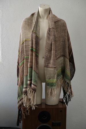 A white painted mannequin stands on a wooden speaker wearing an etherial handwoven scarf in greens and browns