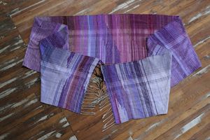 A handwoven length of fabric, woven in every shade of purple is laid out on a wooden floor