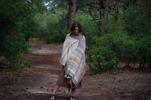 A woman stands on a trail in the forest wearing a handwoven shawl in subtle naturally dyed colors