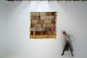 a large blanket, handwoven in bands of naturally dyed colors, hangs on a white gallery wall. a blurry image of a woman moves next to it