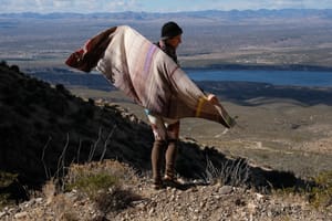 A woman stands on a mountain in a vast desert landscape wearing a handwoven shawl