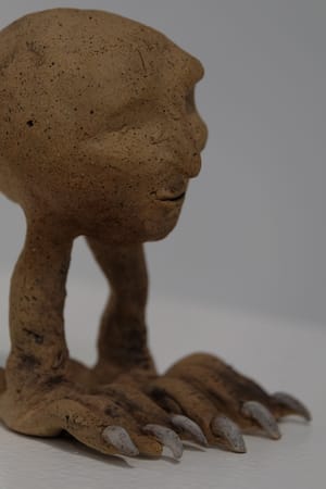 A small clay sculpture of a human head with raven legs. The clay is tan with dark speckles